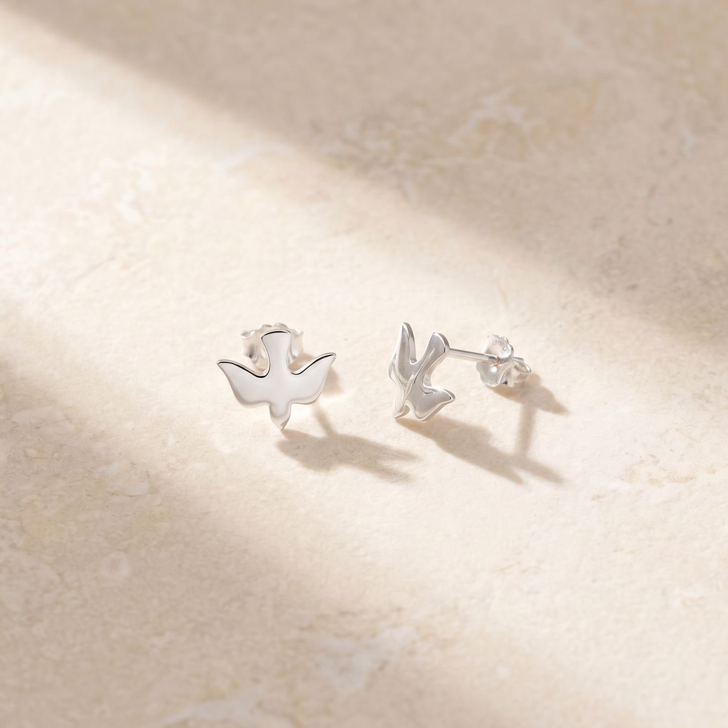 A pair of small silver stud earrings shaped like a swan and a rabbit, displayed on a textured beige surface with soft diagonal shadows.